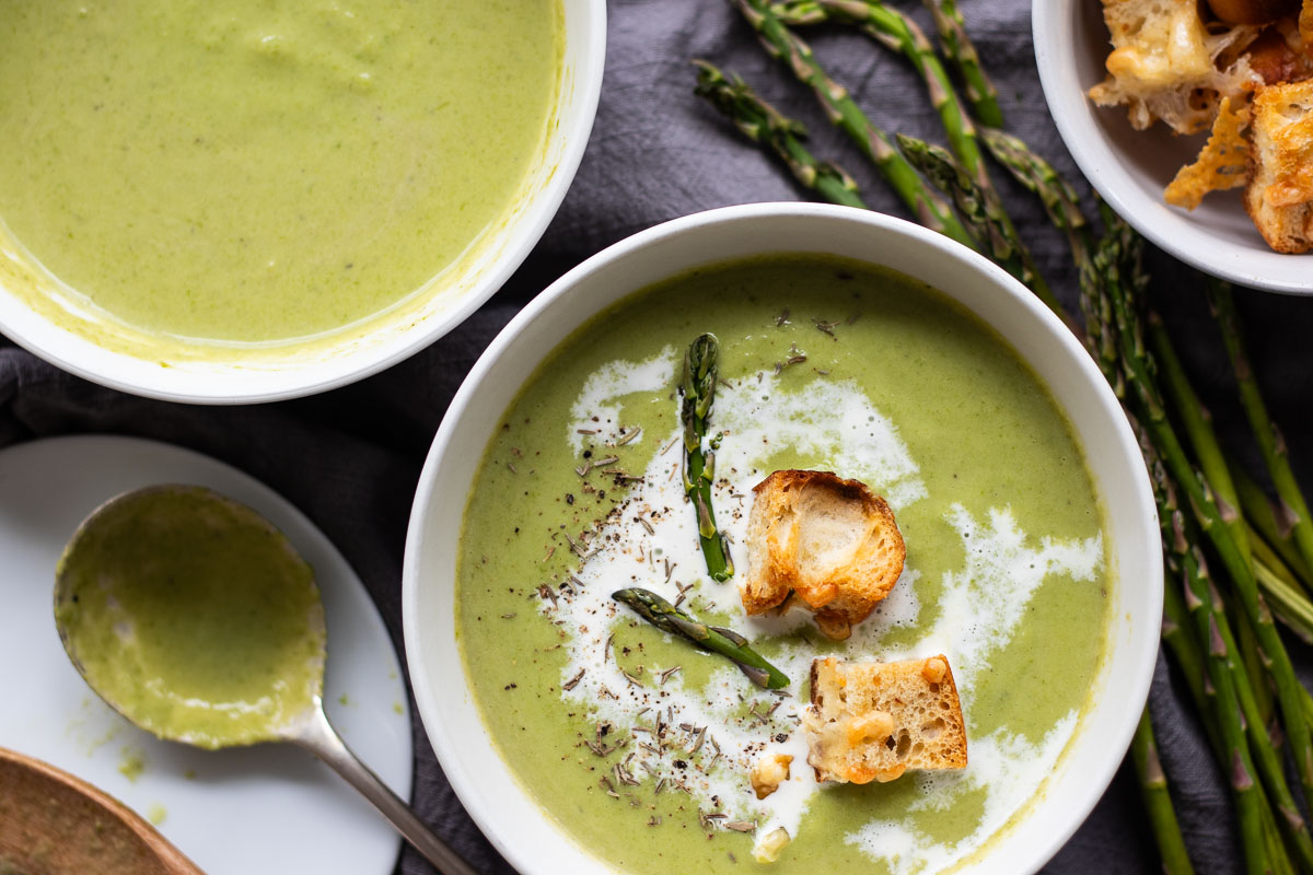 A bowl of creamy asparagus soup garnished with croutons and drizzled with cream. Nearby are fresh asparagus stalks and a spoon with more soup. Another bowl of soup and several croutons are seen in the background.