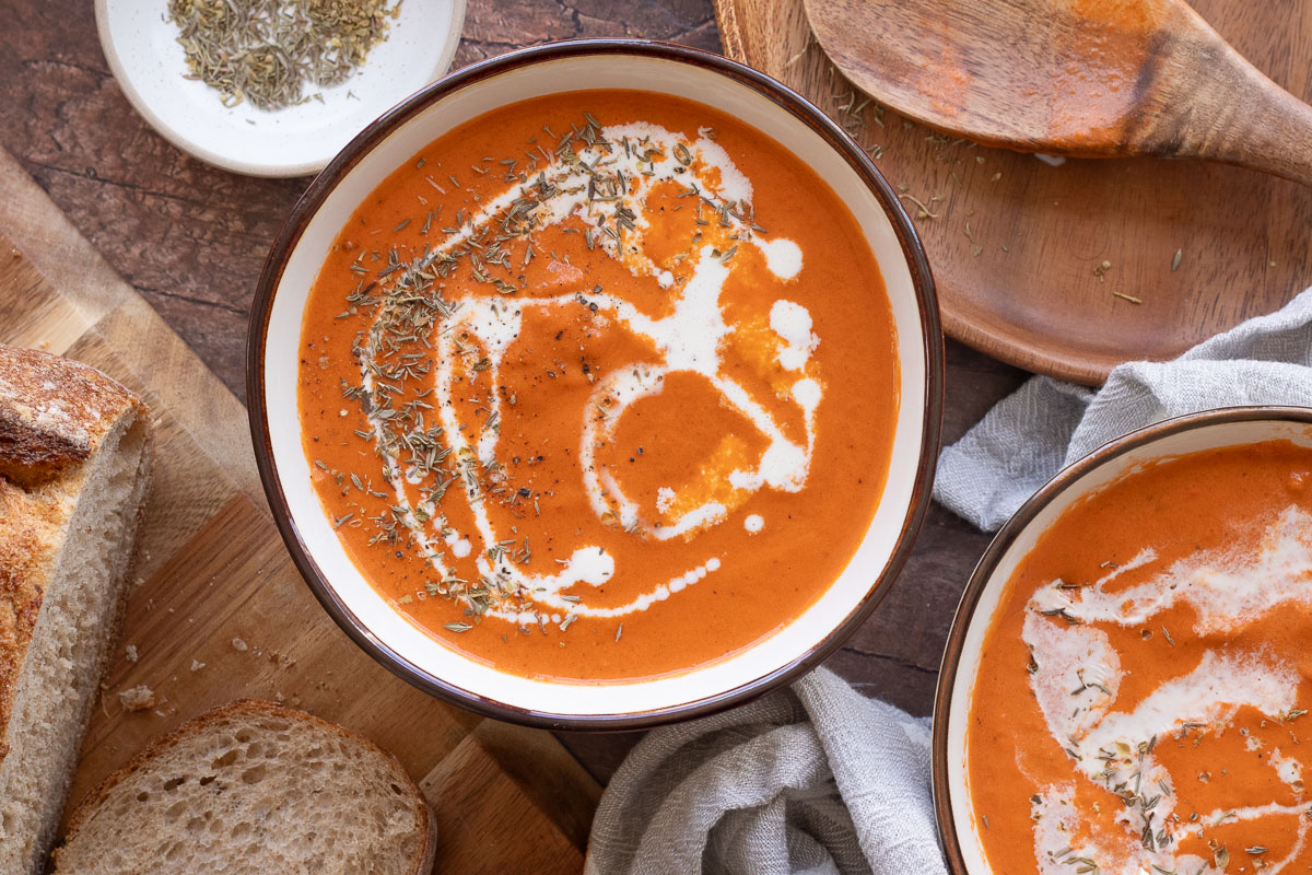 A bowl of creamy tomato soup garnished with a swirl of cream and sprinkled herbs. It's placed on a wooden table with slices of crusty bread nearby. A cloth napkin and a wooden spoon are also visible.