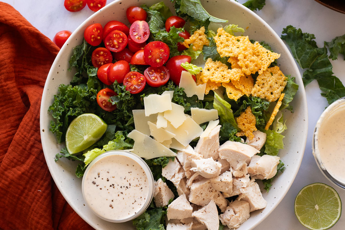 A white bowl with a sweetgreen kale caesar salad copycat. The bowl has kale, lettuce, chicken, tomatoes, parmesan, parmesan crisps, dressing and lime wedge. A red napkin sits nearby as well as small amounts of the ingredients in the bowl.