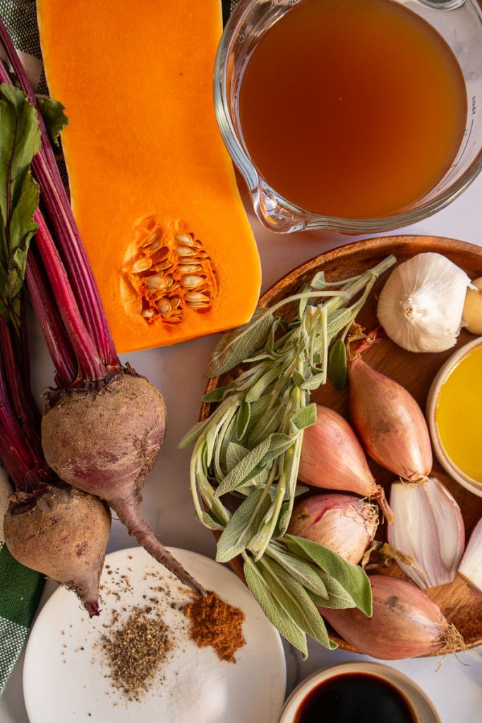 The ingredients for swirled beet and butternut squash soup. There is half of a butternut squash, some fresh beets, vegetable stock, shallots, garlic, sage, balsamic, olive oil, salt pepper and nutmeg.
