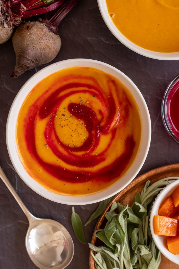 A bowl of beet and butternut squash soup. Some beets sit nearby, as does a spoon and a small bowl of roasted squash and some fresh sage leaves. There is another bowl with some plain butternut squash soup in the top right corner of the image.