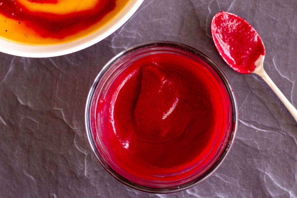 A small clear bowl with purred beets. A spoon with some beets on it sits to the right. In the top left corner of the image there is a corner of a white bowl with some butternut squash and beet soup.