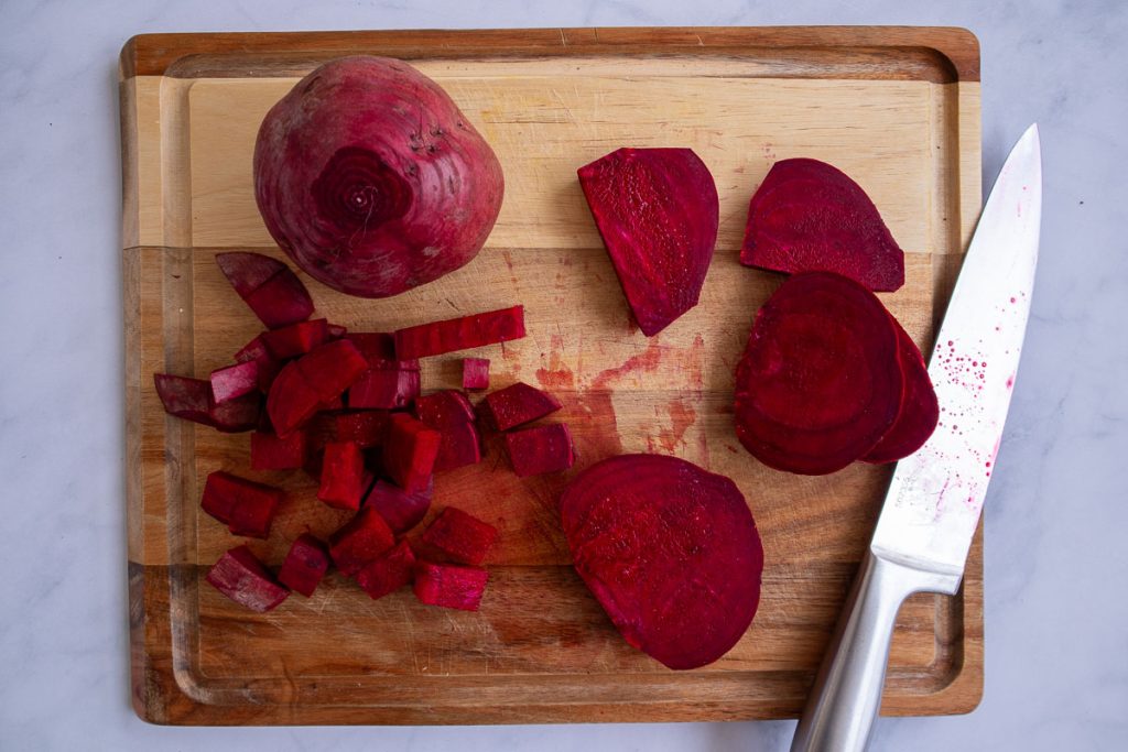A wooden cutting board with some cut and uncut beets and a large chef's knife.
