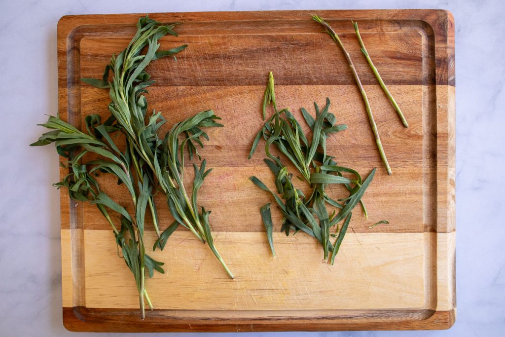 A wooden cutting board with a brunch of fresh tarragon, there is a pile of fresh tarragon leave and some stems sitting next to it.