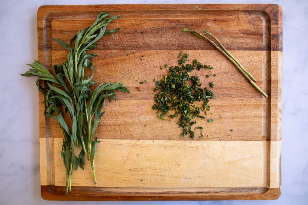 Wooden cutting board with a bunch of fresh tarragon, some chopped tarragon leaves and the stems.