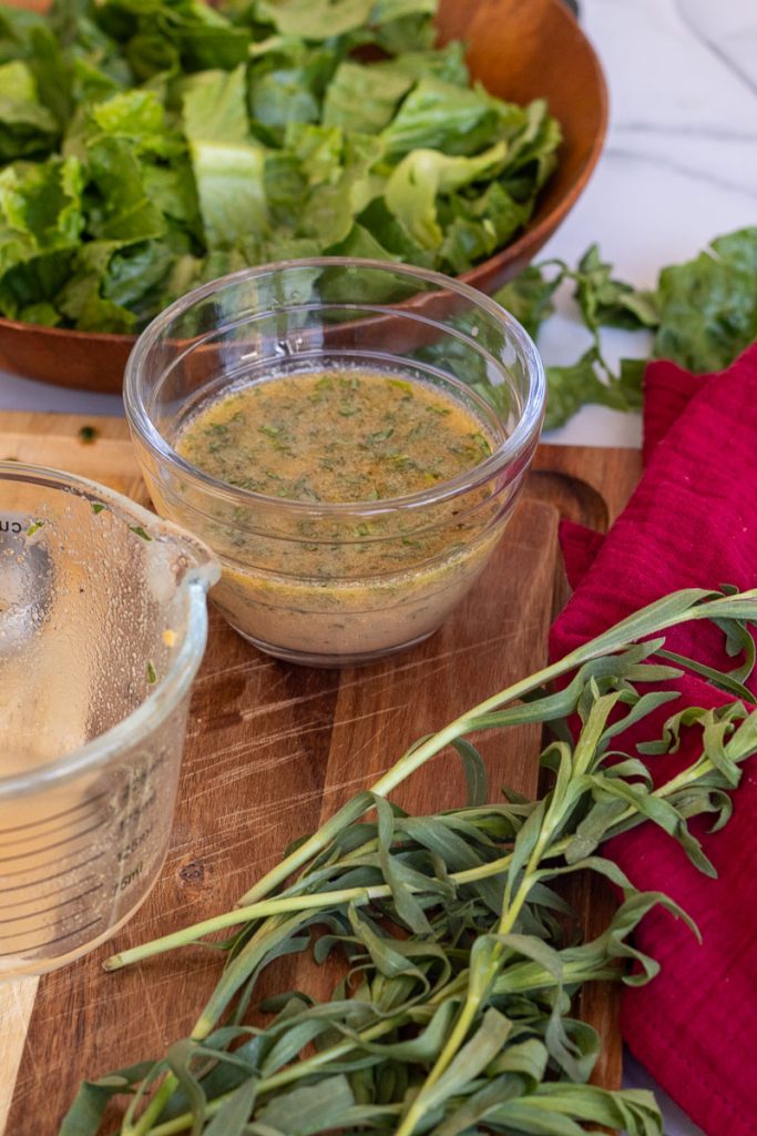 A cutting board with a small container of tarragon vinaigrette on top. There is a red cloth with some fresh tarragon on top and a bowl with lettuce nearby.