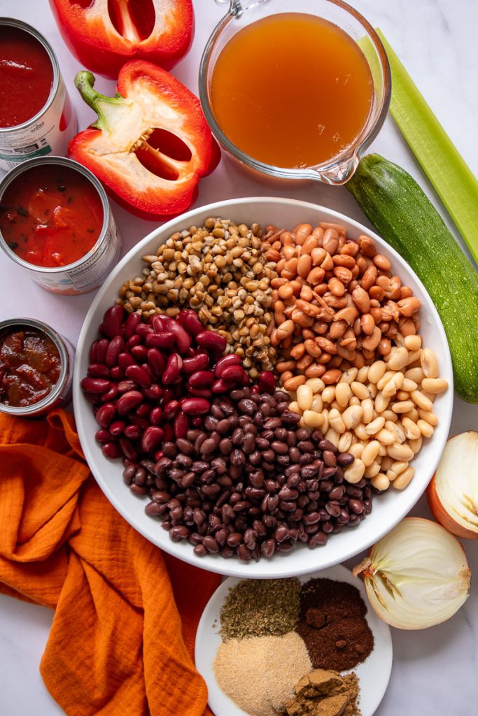 The ingredients for 5-bean chili sitting on a table including a large plate with the five different types of beans, cans of tomato products, spiced, chipotles in adobo, bell pepper, onion, zucchini, celery and vegetable stock.
