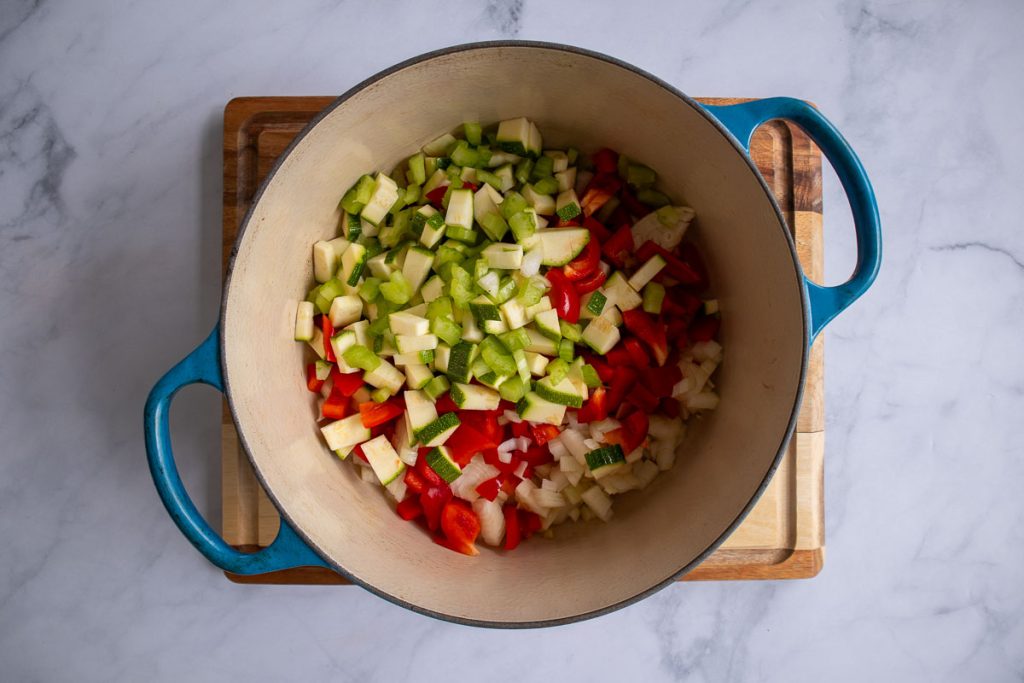 A blue soup pot with uncooked onion, celery, bell pepper and zucchini.