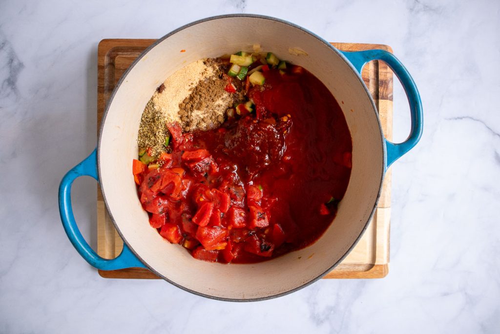 A blue soup pot with cooked onion, celery, bell pepper and zucchini, as well as spices, canned tomatoes and chipotles unmixed.