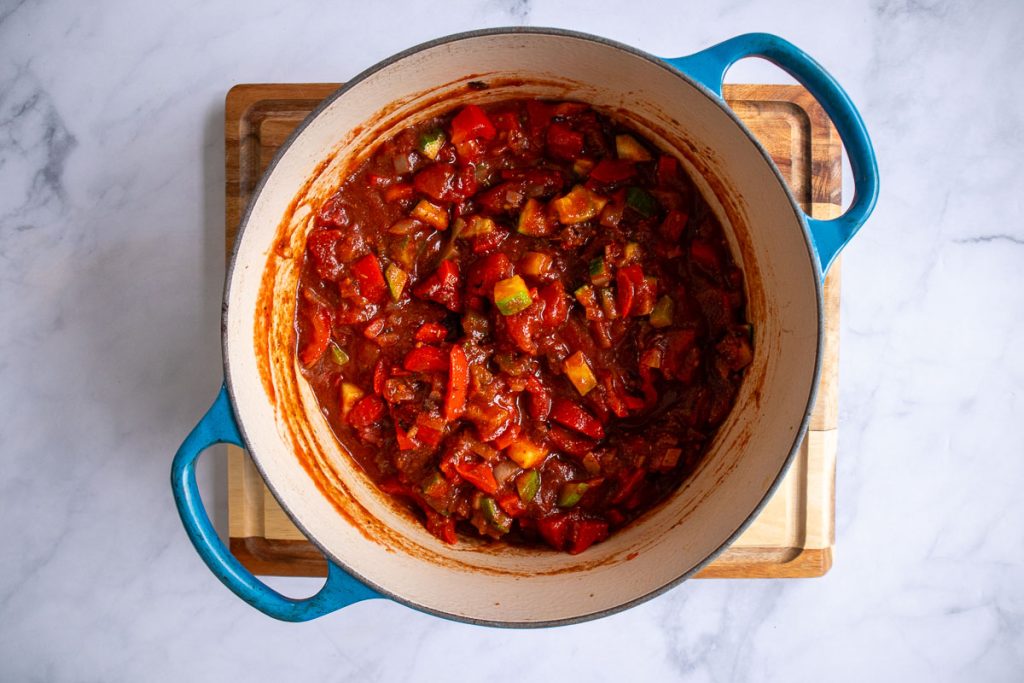 A blue soup pot with cooked onion, celery, bell pepper and zucchini, as well as spices, canned tomatoes and chipotles mixed.