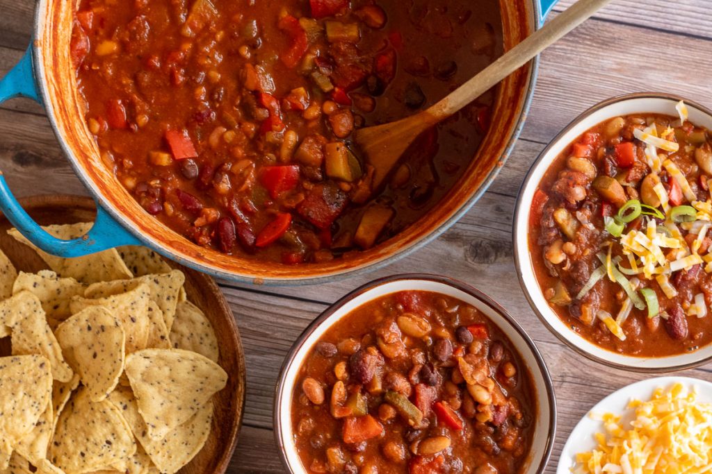 A large blue soup pot with 5-bean chili and a wooden spoon in it. There are two bowls with chili sitting nearby as well as some toppings like tortilla chips and shredded cheese.