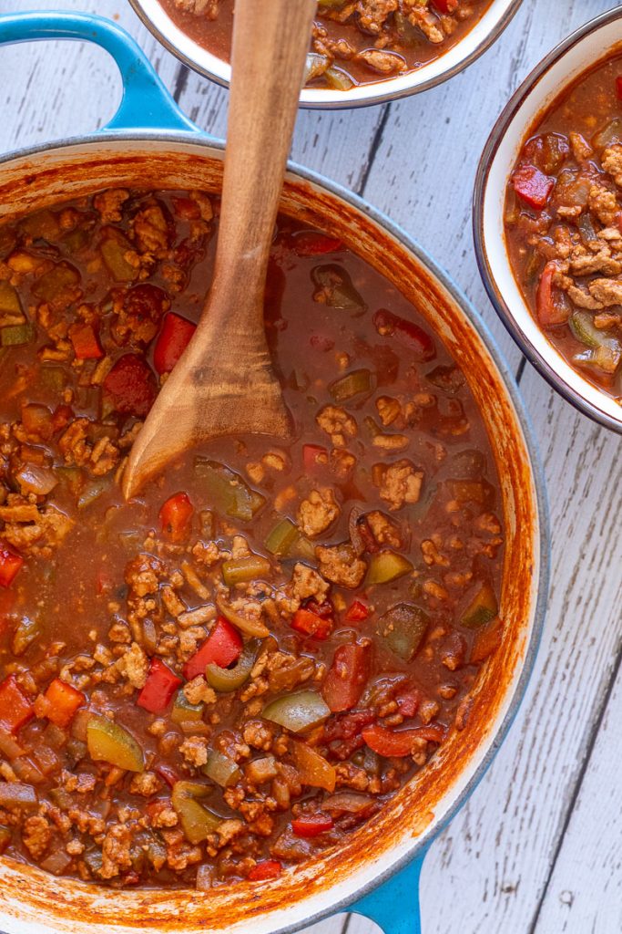 A large blue soup pot with cooked turkey vegetable chili. There is a wooden spoon in it and some smaller bowls filled with chili nearby.