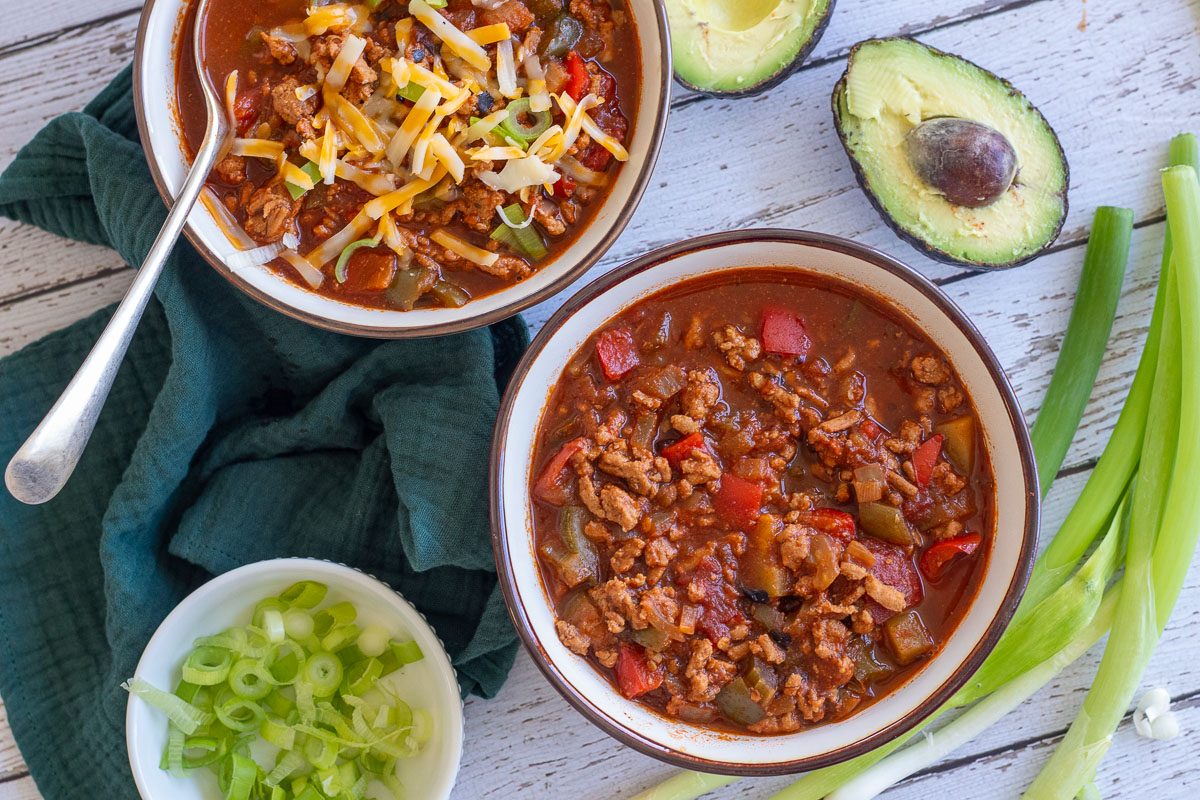Two bowls of turkey vegetable chili sit on a table. There are two halves of an avocado, some green onions, sliced green onions in a little white bowl and a green cloth nearby.