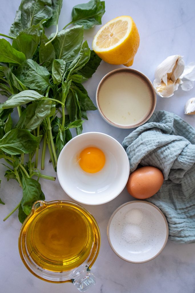 Ingredients for basil mayonnaise on a table. There is a large bunch of basil, lemon, avocado oil, two eggs, salt, sugar and garlic.