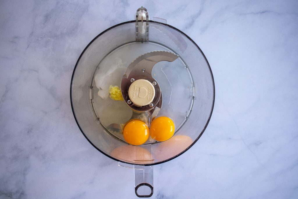 An overhead shot of a food processor bowl with raw eggs, lemon juice, garlic, salt and sugar, unprocessed.