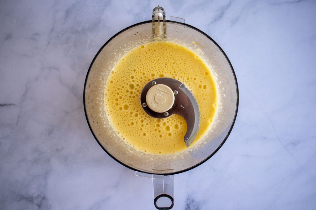 An overhead shot of a food processor bowl with eggs, lemon juice, garlic, salt and sugar processed.