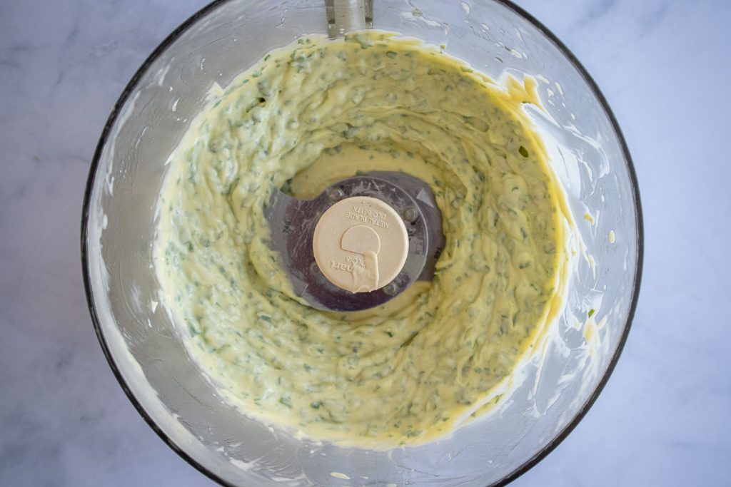A close up overhead shot of a food processor bowl with homemade basil mayonnaise inside.