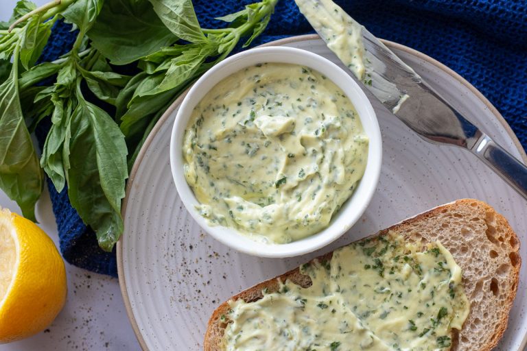 A white plate with a small white bowl with basil mayo in it. There is a knife and a piece of bread on the plate. The bread has mayo on it. There is also a knife on the plate and some basil and a lemon off to the side.