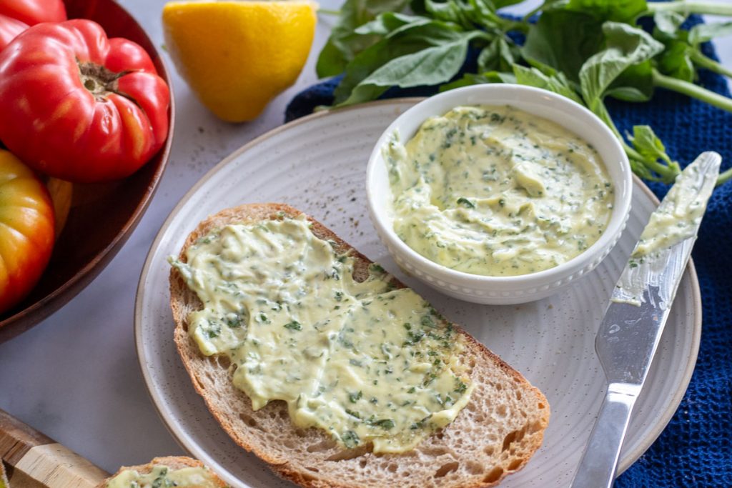A plate with a piece of bread slathered in basil mayonnaise. There is a small bowl on the plate with more mayo and a knife as well. There is some basil in the top right and a bowl of tomatoes in the top left of the image.