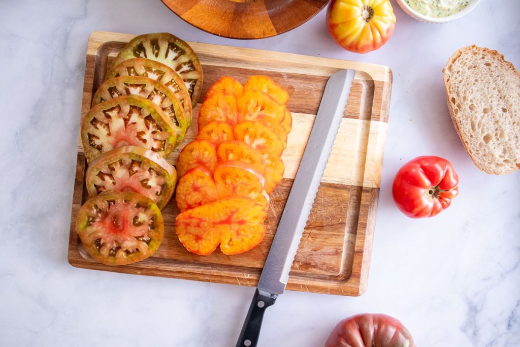 A wooden cutting board with some sliced heirloom tomatoes and a serrated knife sitting on top. There are some uncut tomatoes and sliced bread nearby.