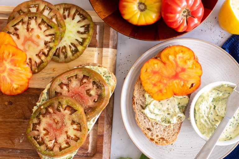 A wooden cutting board with a piece of tomato toast and some sliced heirloom tomatoes nearby. There is a bowl with uncut tomatoes at the top and a white plate with a piece of toast in the process of being assembled. There is a bowl of basil mayo and a knife nearby.